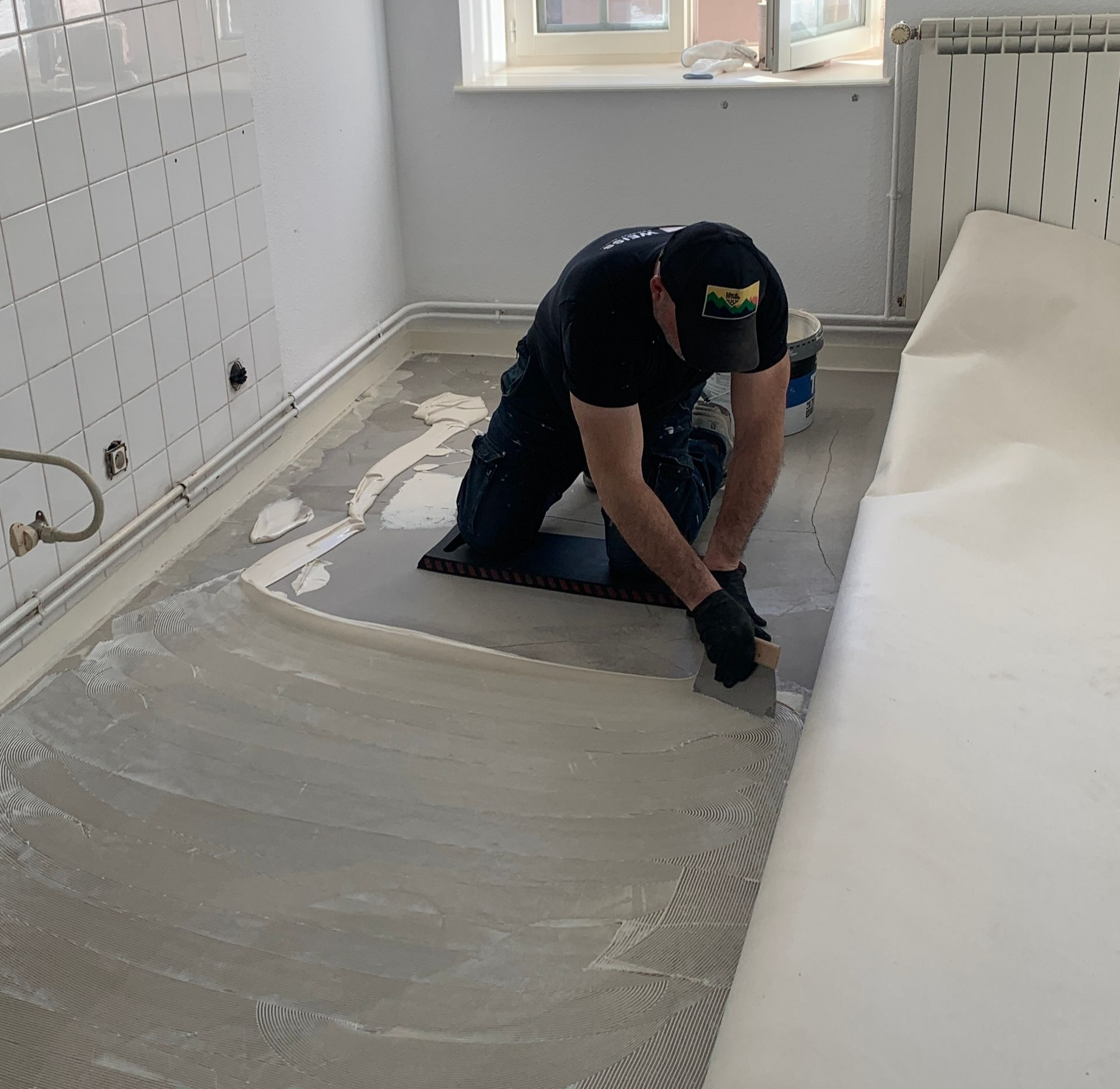 Un homme agenouillé, posant un revêtement de sol à la truelle, dans un couloir.