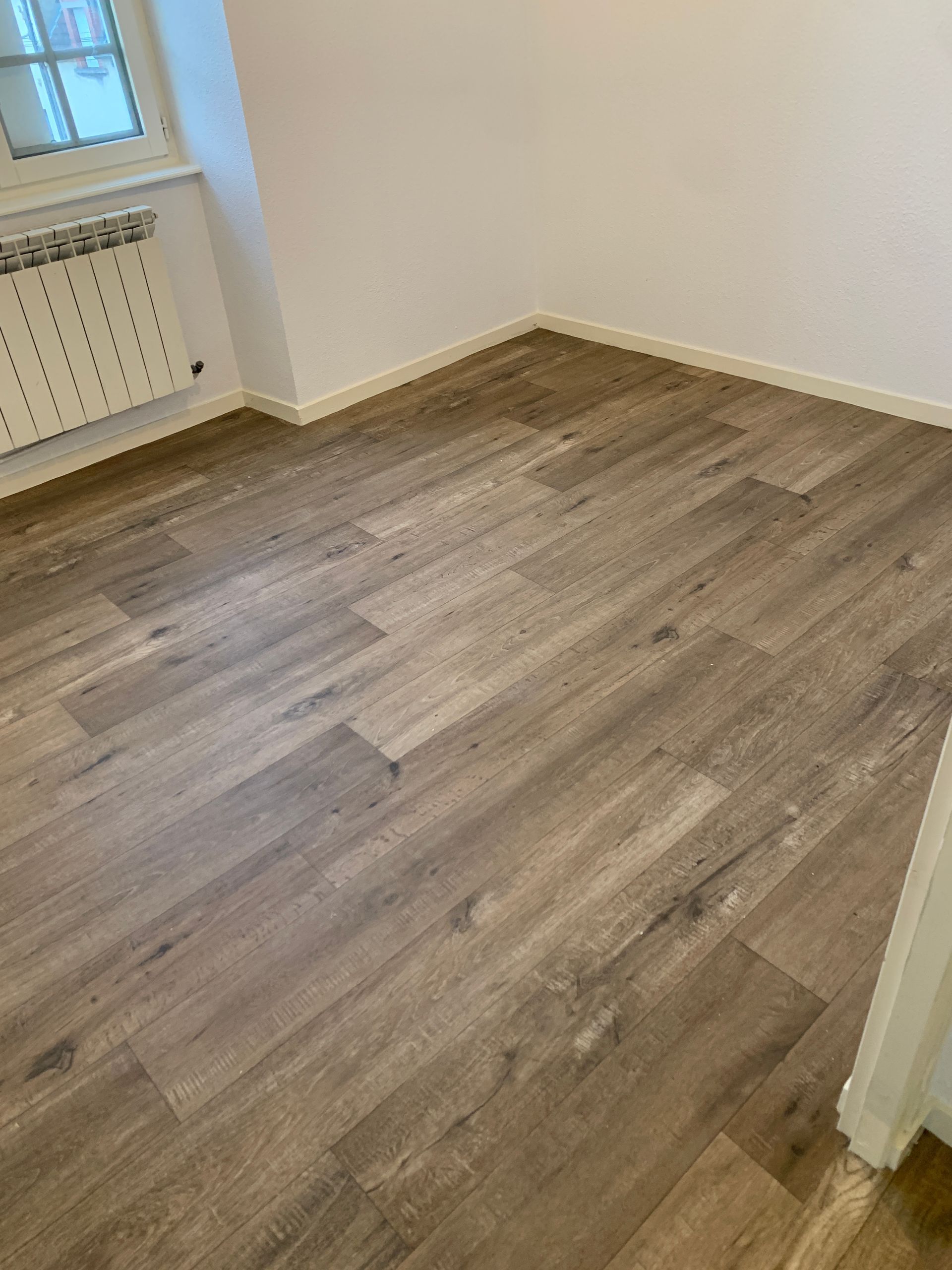 Pièce vide avec sol imitation bois et murs blancs.