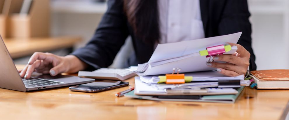 Une personne utilise un ordinateur portable à côté d'une pile de papiers sur un bureau.