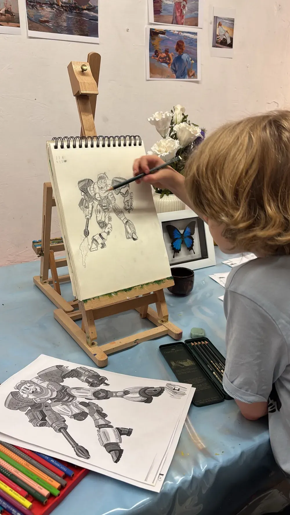 Una persona dibuja en un caballete en un estudio de arte, con dibujos terminados y lápices de colores cerca.