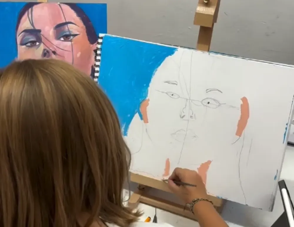 Una persona pinta un rostro sobre un lienzo en un estudio de arte, con un retrato terminado detrás.