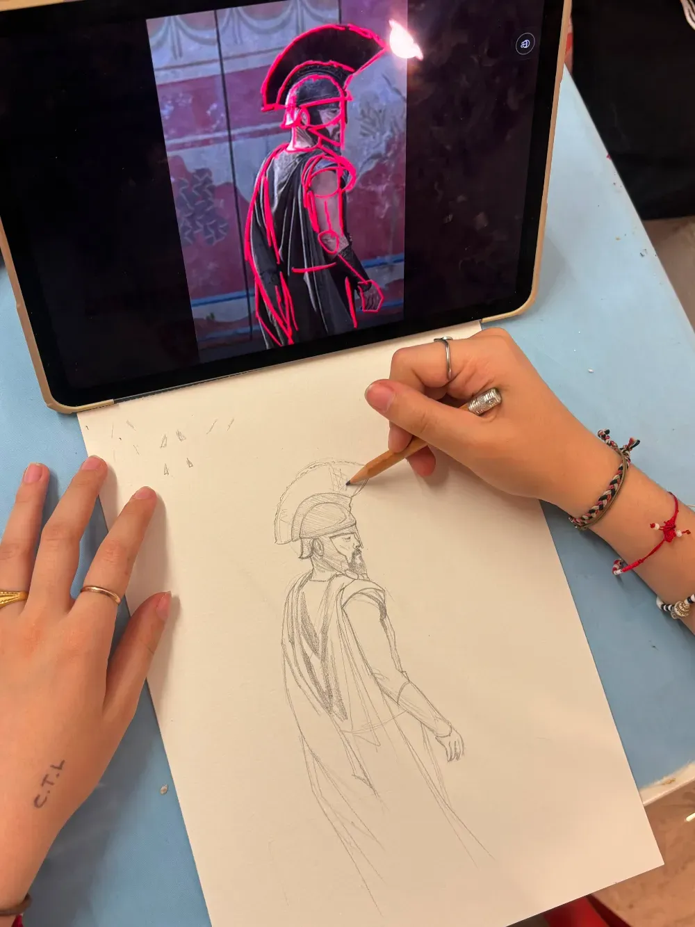 Manos dibujando una figura a partir de una referencia de color rojo neón en una tableta, sobre un dibujo a lápiz en papel.