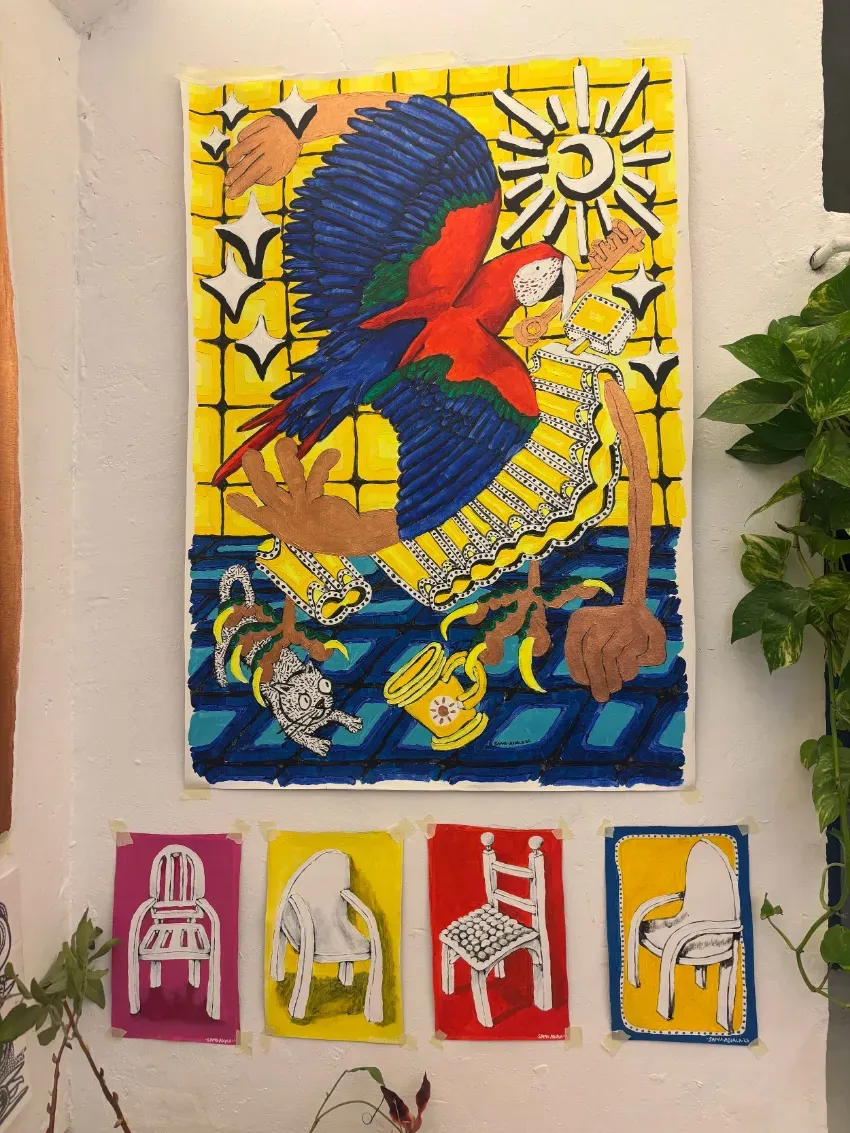 Un mural colorido de loros en una pared, con cuatro pequeños paneles de sillas pintadas debajo, junto a una planta.