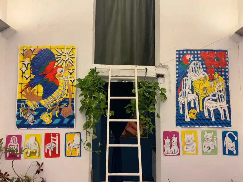 Pared de galería colorida con dos grandes pinturas abstractas, una escalera, plantas colgantes y pequeños grabados enmarcados debajo.