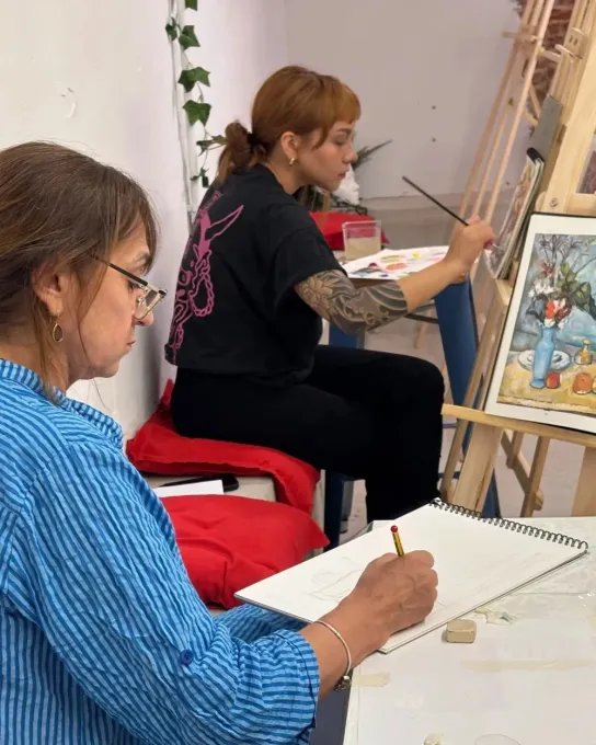 Dos personas dibujando en caballetes en un estudio de arte, una vestida de azul y la otra de negro, con una silla roja.