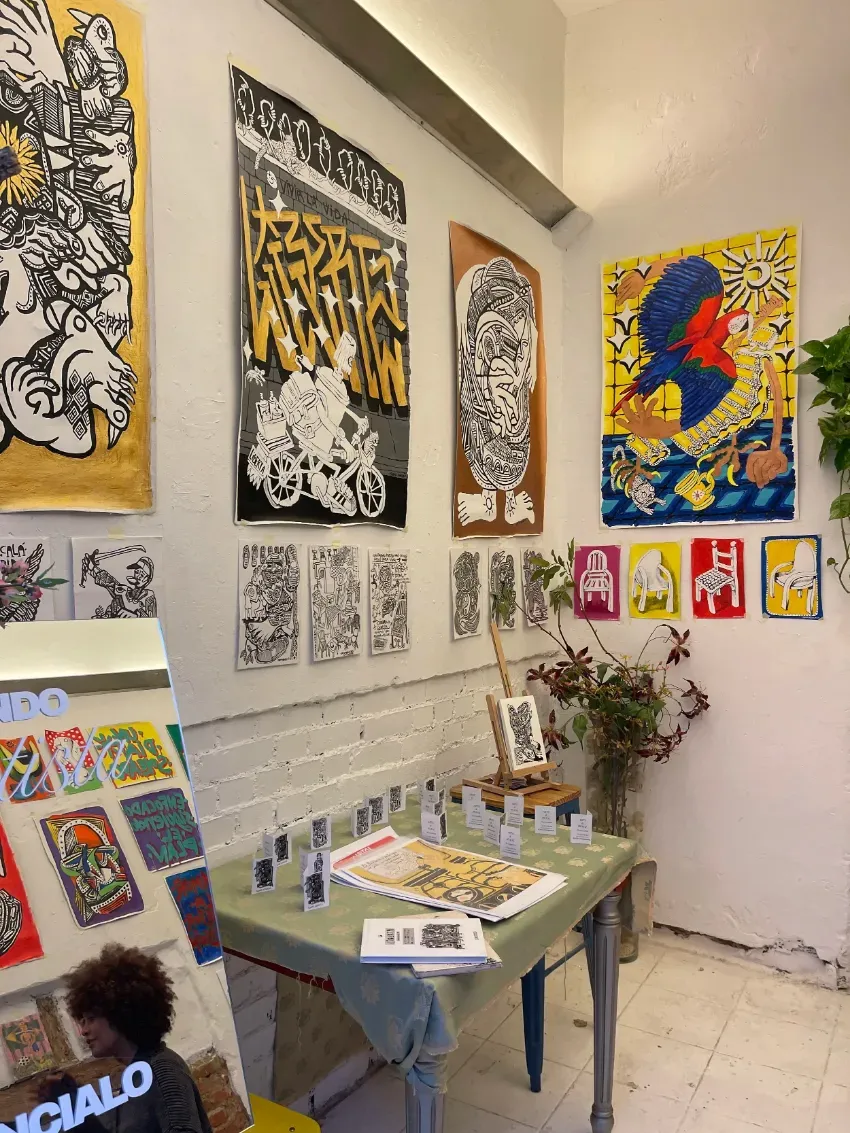 Pequeño estudio de arte con coloridos carteles, láminas en la pared y una mesa con materiales de arte y tarjetas.