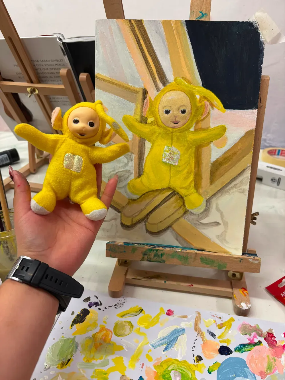 Una mano sostiene un peluche amarillo de Teletubby junto a un lienzo pintado a juego sobre un caballete.