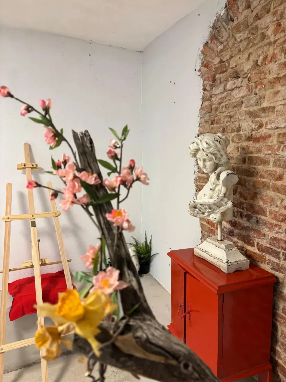 Armario rojo con una escultura de busto blanco contra una pared de ladrillos, con una rama florida en primer plano.
