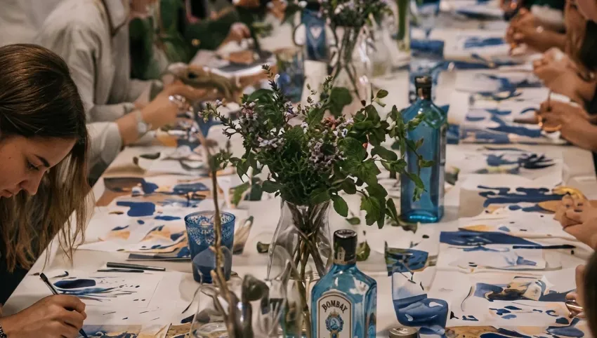 Mesa larga de comedor preparada para una comida, con botellas de vidrio azul, flores y los invitados sentados a ambos lados.