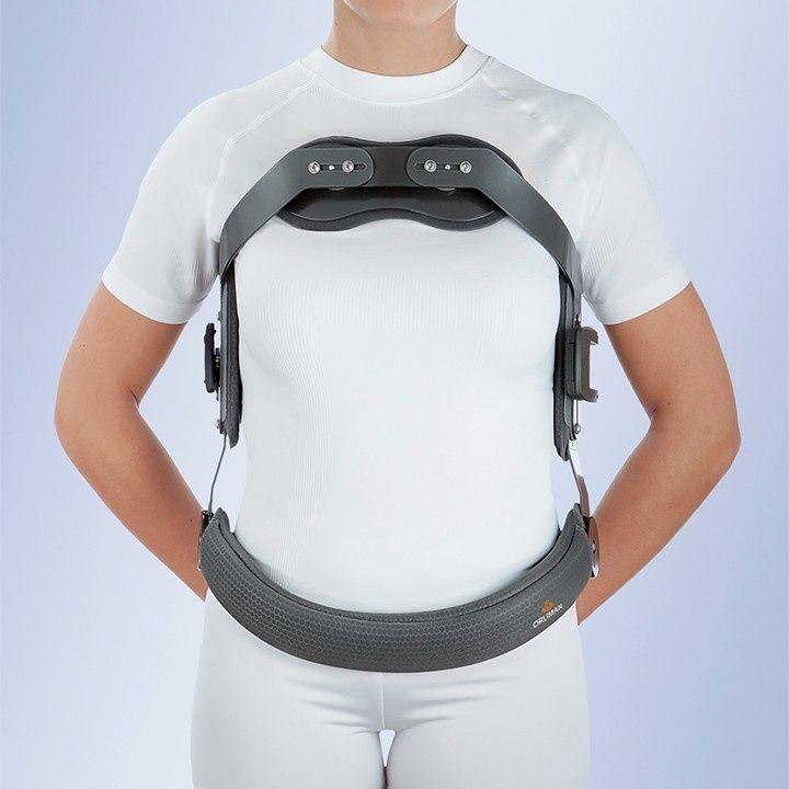 Persona que lleva un corsé ortopédico gris sobre una camisa y pantalones blancos, de pie con las manos en las caderas.