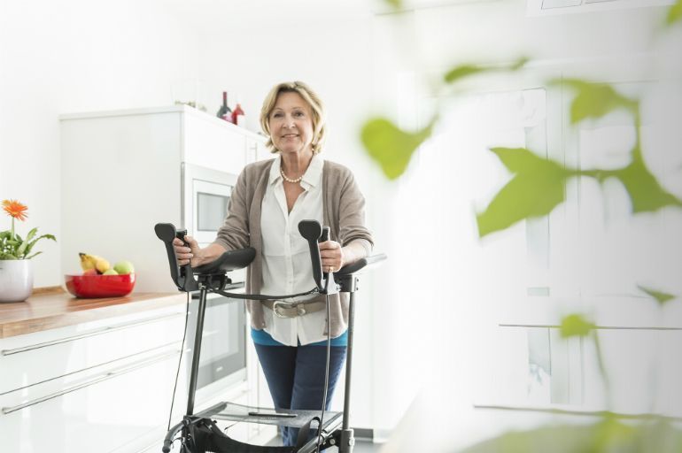 Mujer que usa un andador en una cocina luminosa, sonriendo junto a una encimera con frutas y plantas.