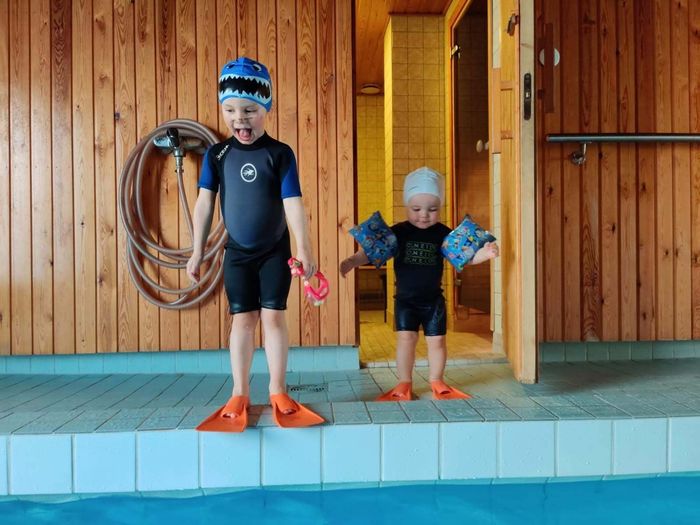 Nuori lapsi uimavarusteissa ja taapero kellukkeissa seisovat uima-altaan reunalla - Helsingin Vauvauimarit Oy