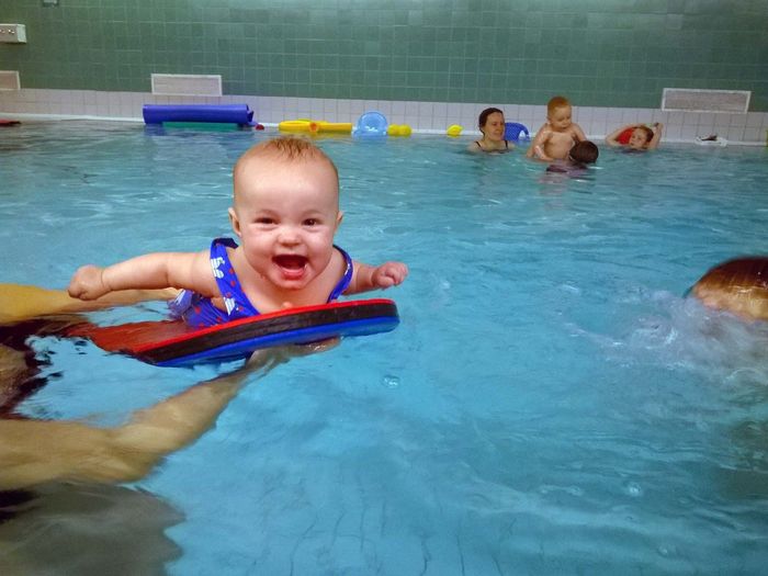 Naurava vauva katsoo suoraan kameraan kelluessaan vedessä uimalaudan päällä - Helsingin Vauvauimarit Oy