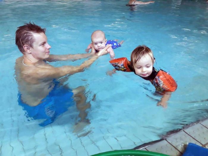 Isä kannattamassa vauvaa vedessä nuoren lapsen kelluessa kellukkeiden avulla vieressä - Helsingin Vauvauimarit Oy