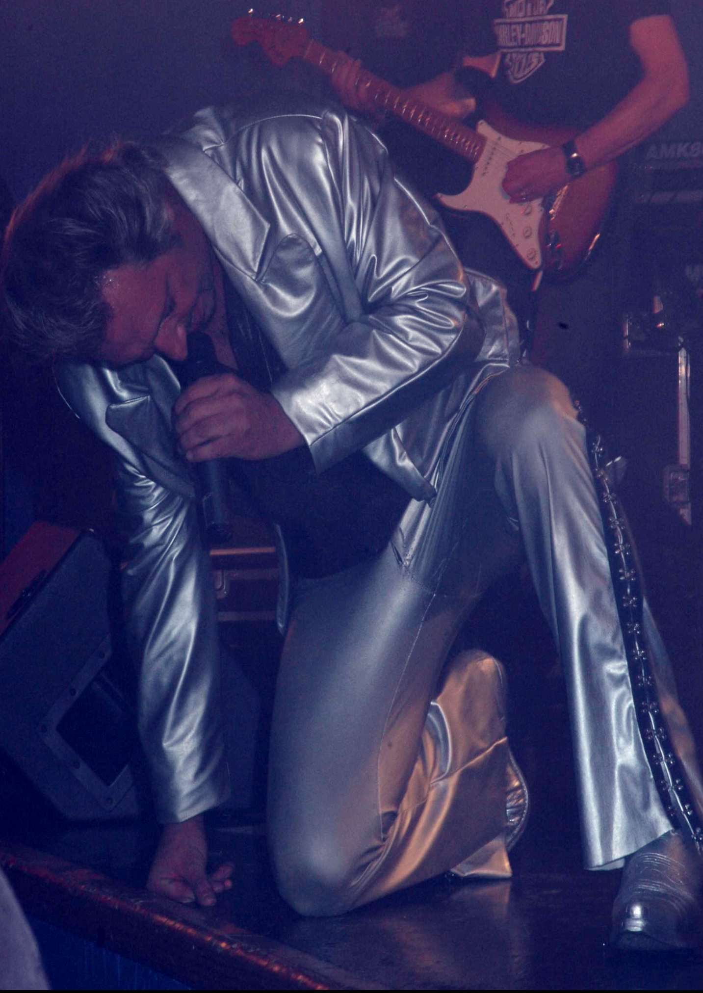 JOHNNY DEMS en costume argenté, agenouillé, chante dans un micro. Un guitariste est en arrière-plan.