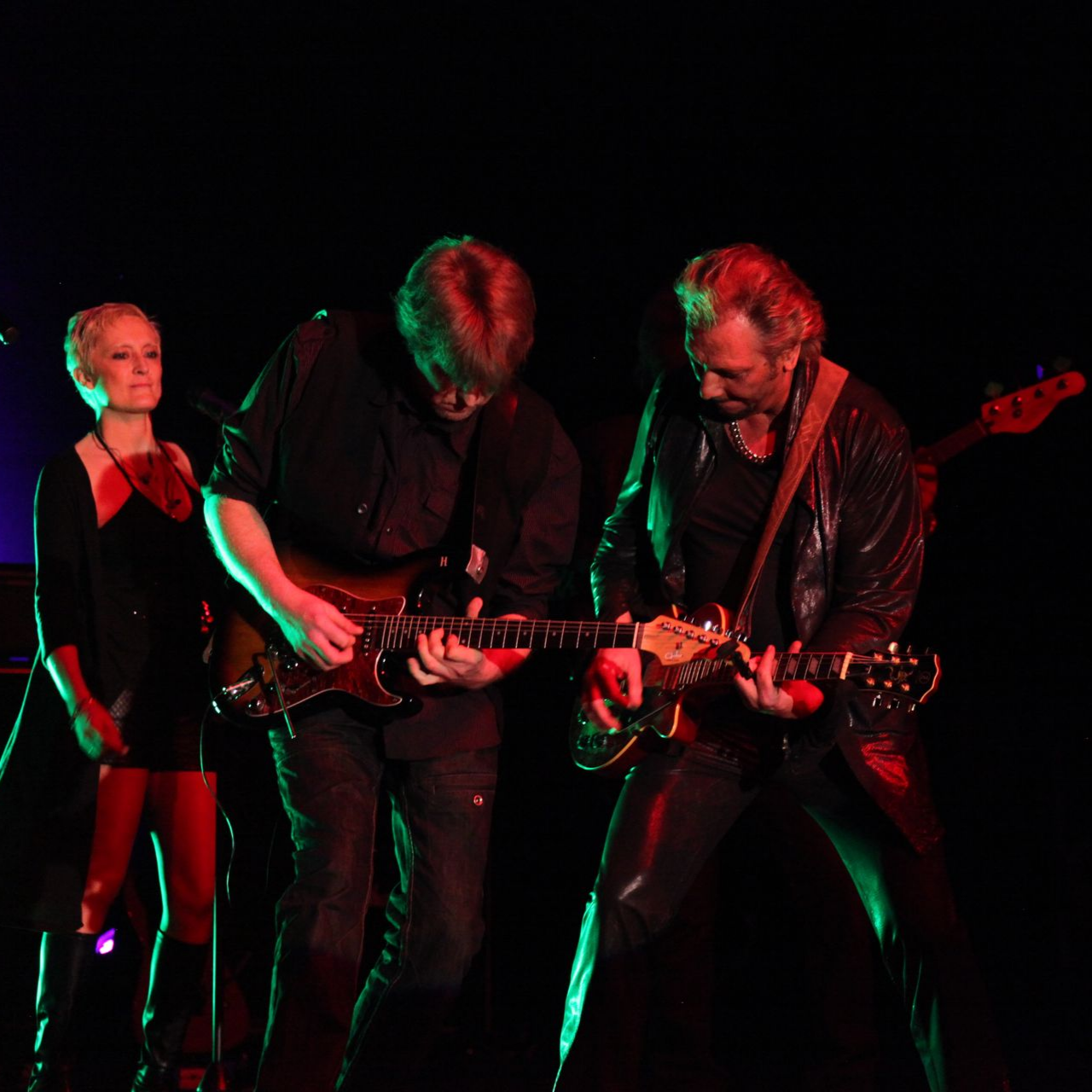 Un groupe joue sur scène, JOHNNY DEMS et un guitariste jouent, une femme se tient à proximité.