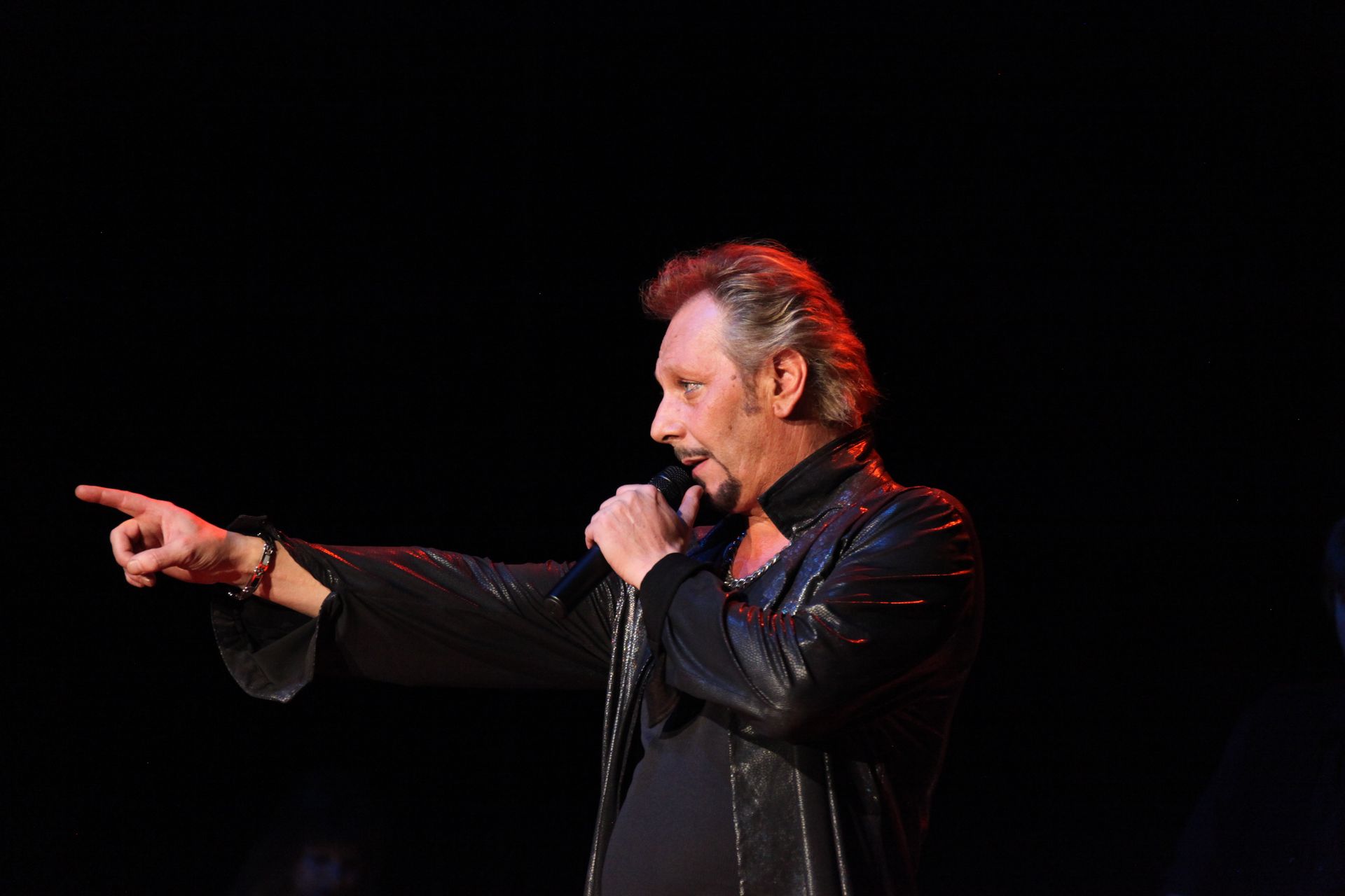 JOHNNY DEMS  chante sur scène, pointant du doigt sur le côté, vêtu de noir, sous un éclairage rouge.