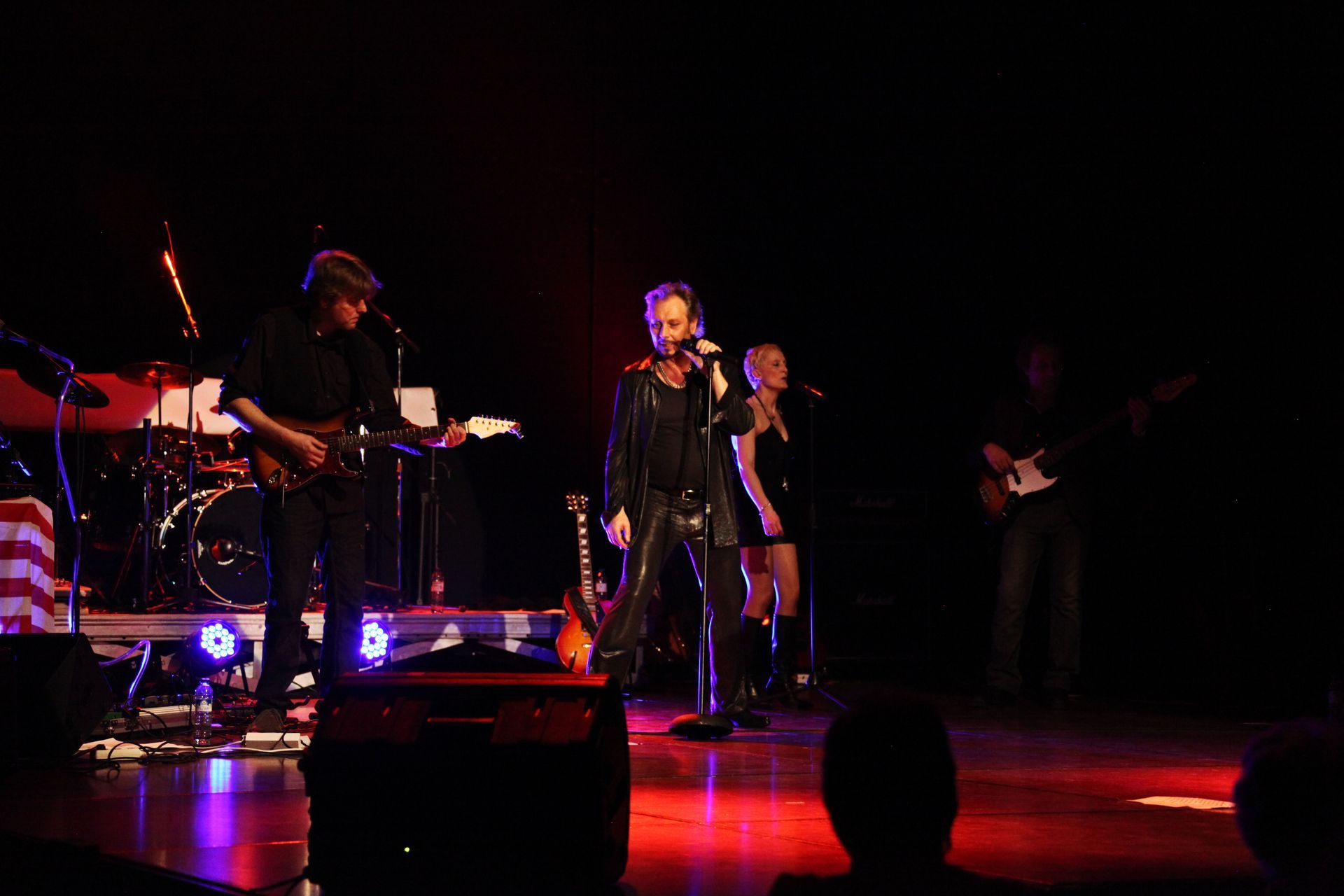 Le groupe se produit sur scène : JOHNNY DEMS en costume de cuir noir, un guitariste, un batteur et une choriste.