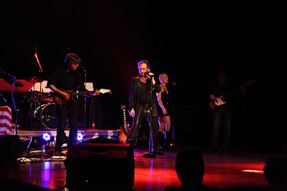 Le groupe se produit sur scène : JOHNNY DEMS en costume de cuir noir, un guitariste, un batteur et une choriste.