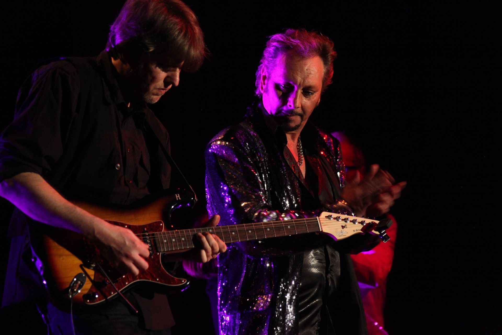 Deux musiciens sur scène, l'un jouant de la guitare et JOHNNY DEMS vêtu d'une veste à paillettes.