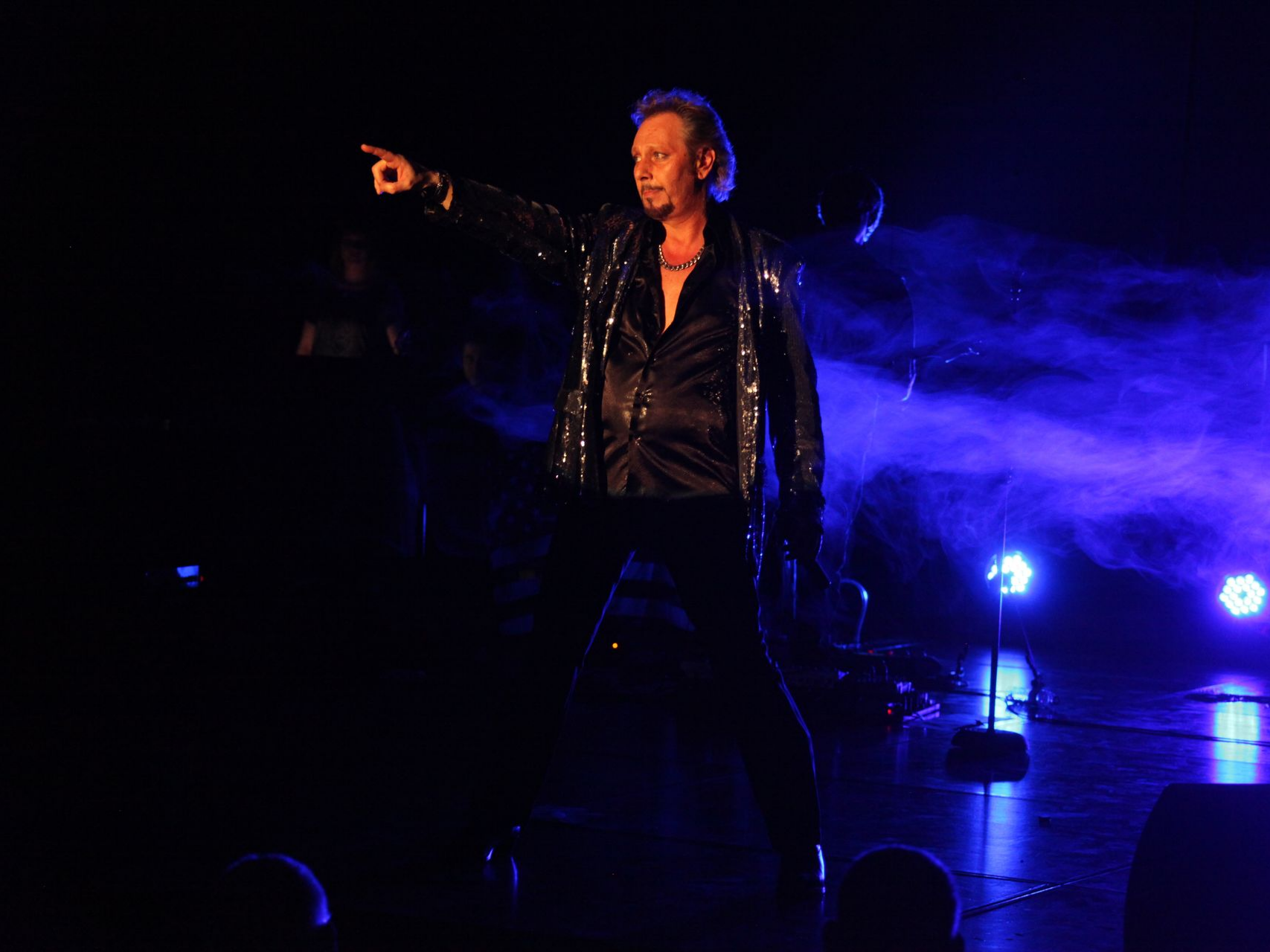 Sur une scène éclairée de bleu, JOHNNY DEMS vêtu d'une veste noire à paillettes pointe du doigt.