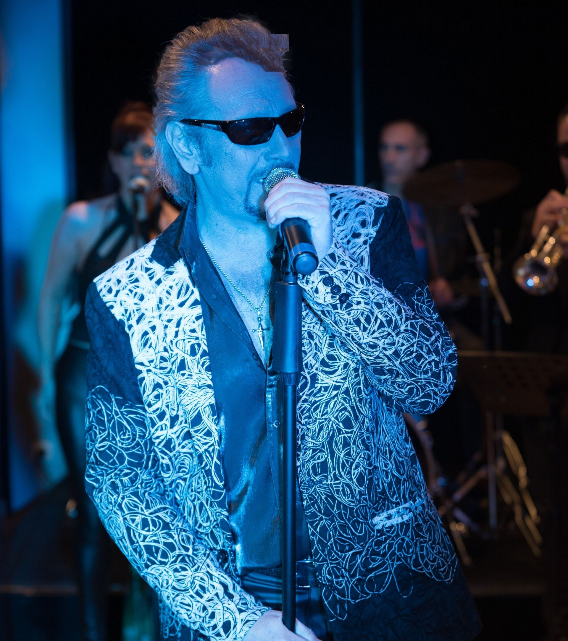 JOHNNY DEMS chante dans un microphone, portant des lunettes de soleil et une veste à motifs sur scène.