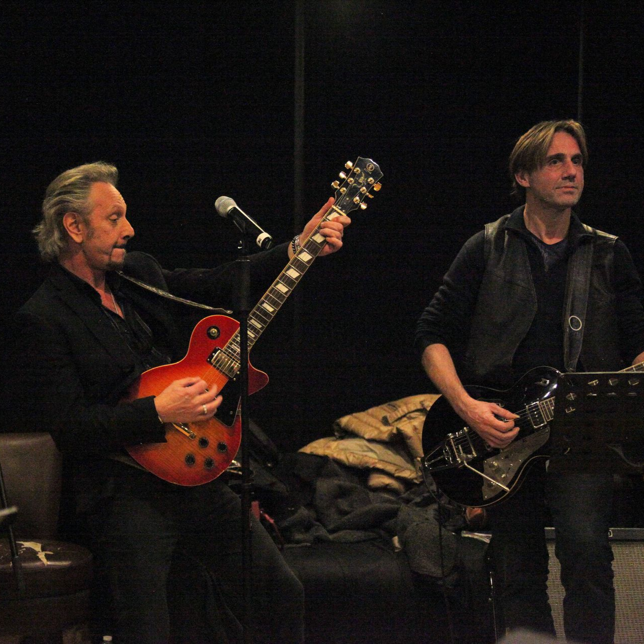 Deux guitaristes jouent sur scène. JOHNNY DEMS  tient une guitare sunburst, l'autre une guitare noire.