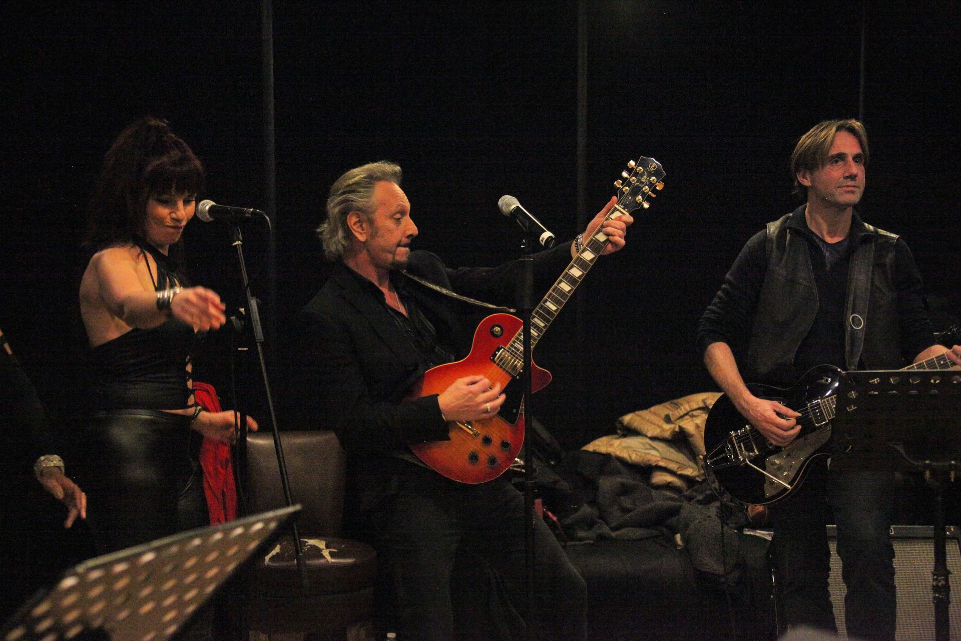 Un groupe se produit sur scène : une femme chante, JOHNNY DEMS tient sa guitare, un autre guitariste joue.