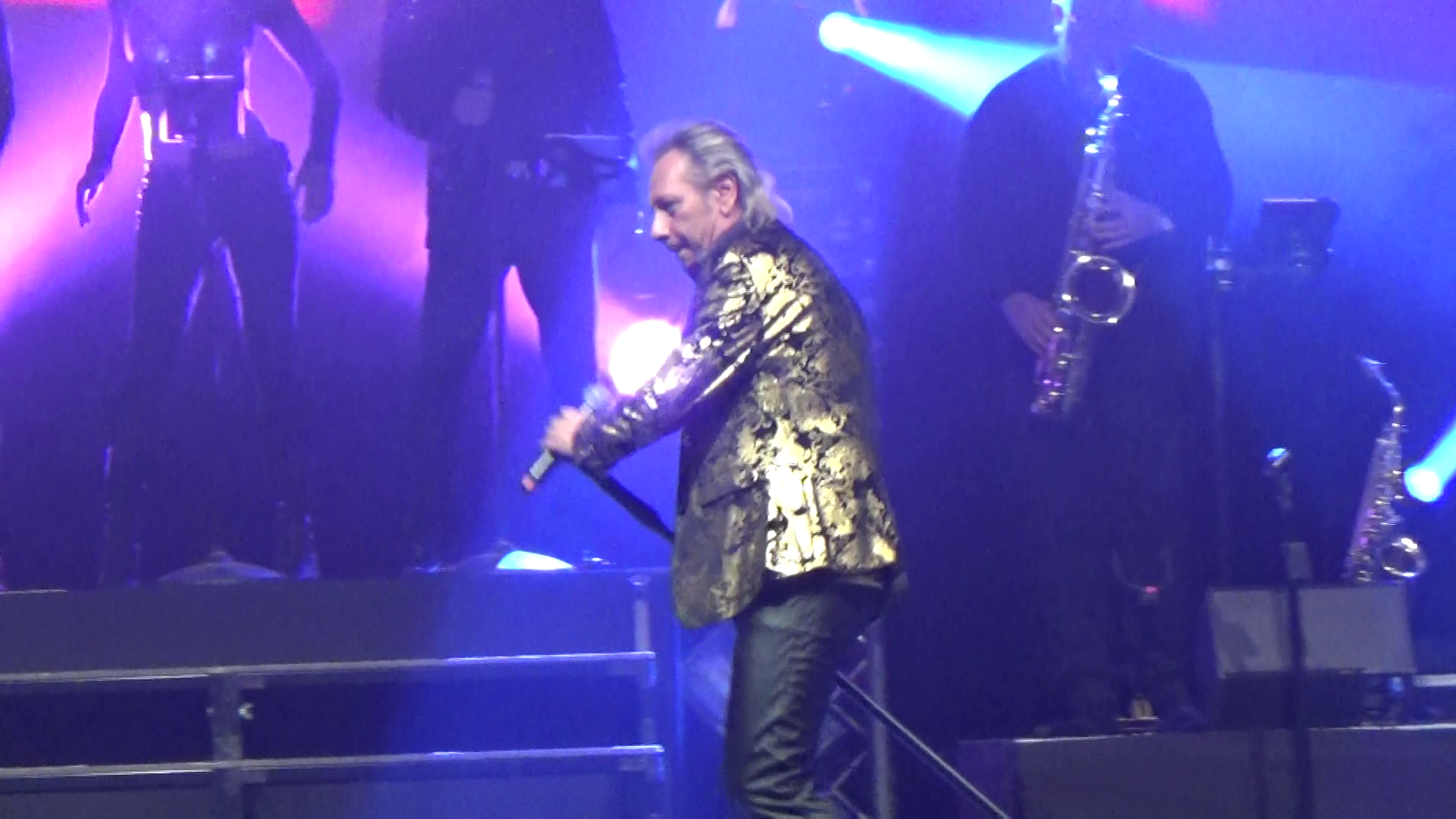 JOHNNY DEMS en veste dorée est sur scène, micro en main. Les membres du groupe sont visibles en arrière-plan.