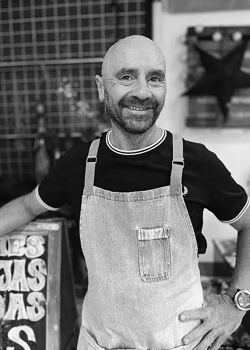 Un hombre vestido con un mono y una camisa negra sonríe en una foto en blanco y negro.
