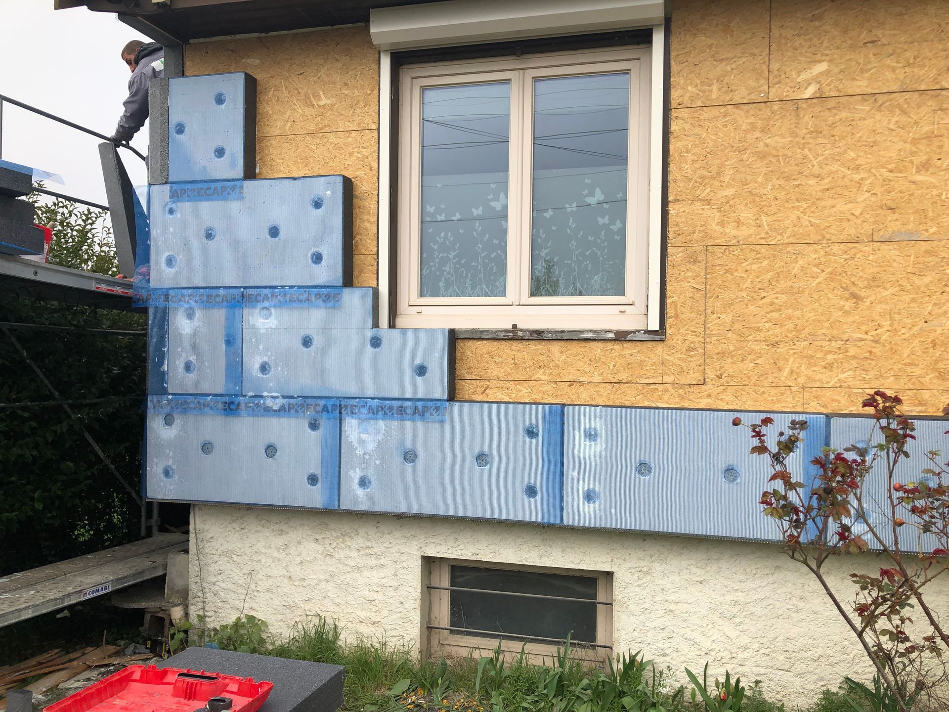 Un homme installe des panneaux isolants bleus sur l'extérieur d'une maison, près d'une fenêtre et d'un échafaudage.