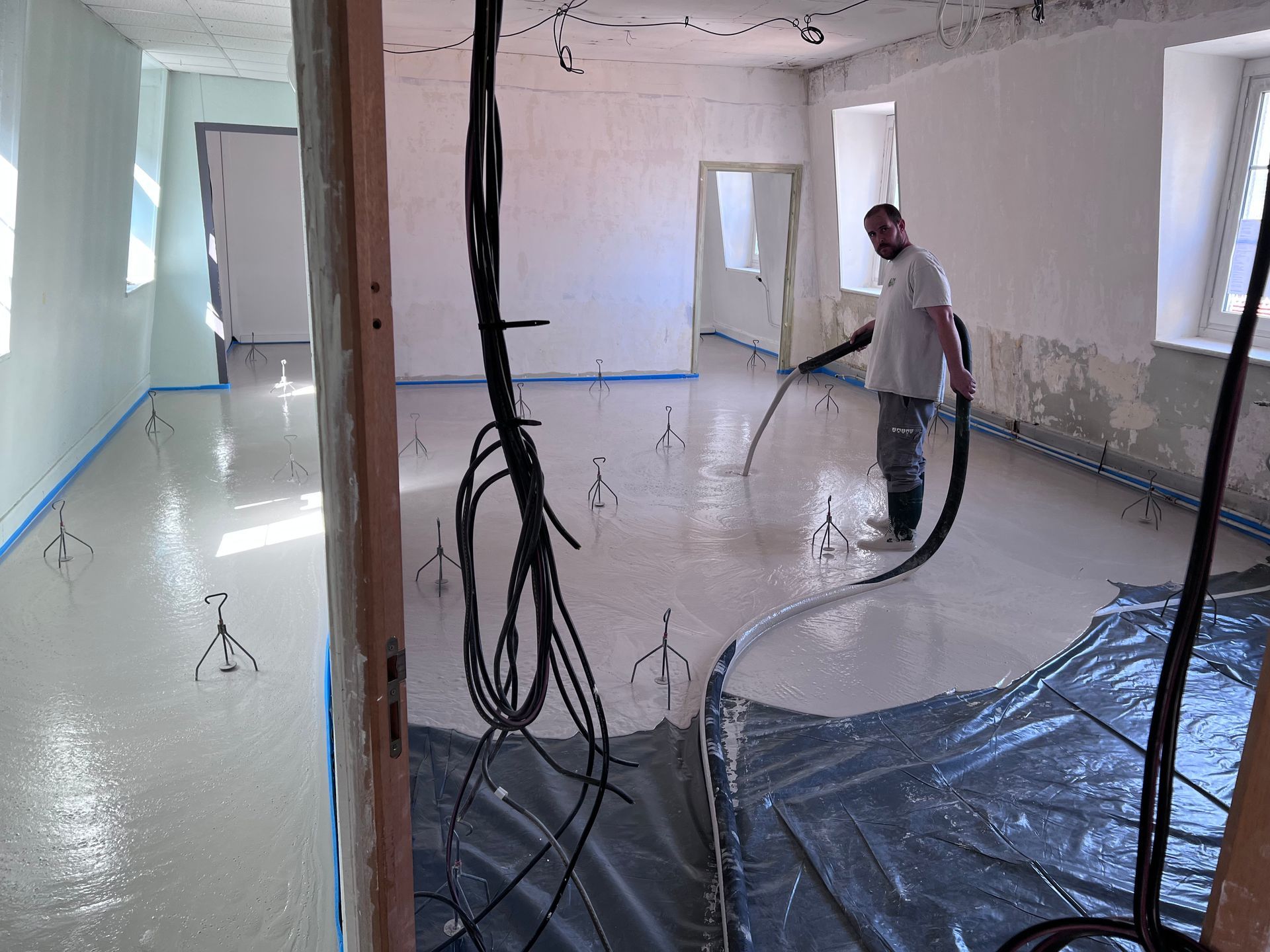 Un homme applique du béton autonivelant sur le sol d'une pièce ; des becs, des fils et du ruban bleu sont visibles.