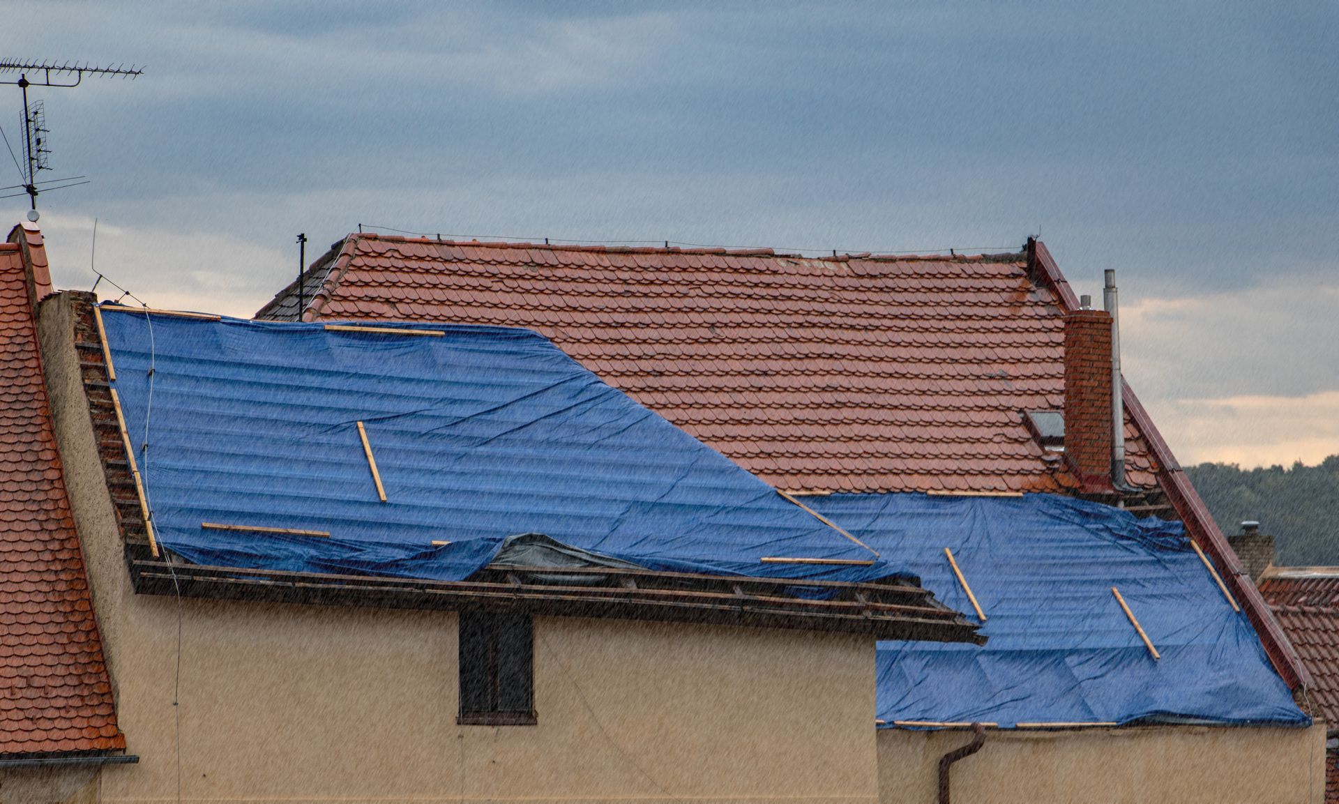 Une bâche bleue recouvre un toit endommagé, contrastant avec les tuiles rouges environnantes.