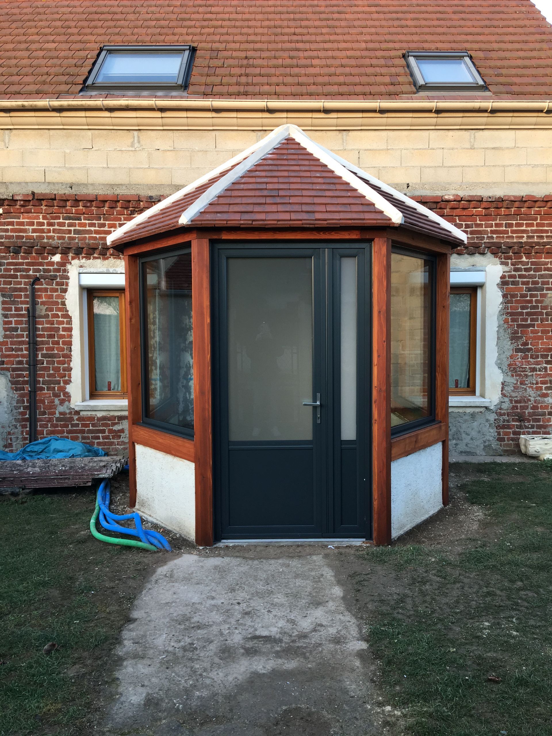 Porche octogonal en bois avec une porte sombre contre un bâtiment en briques.