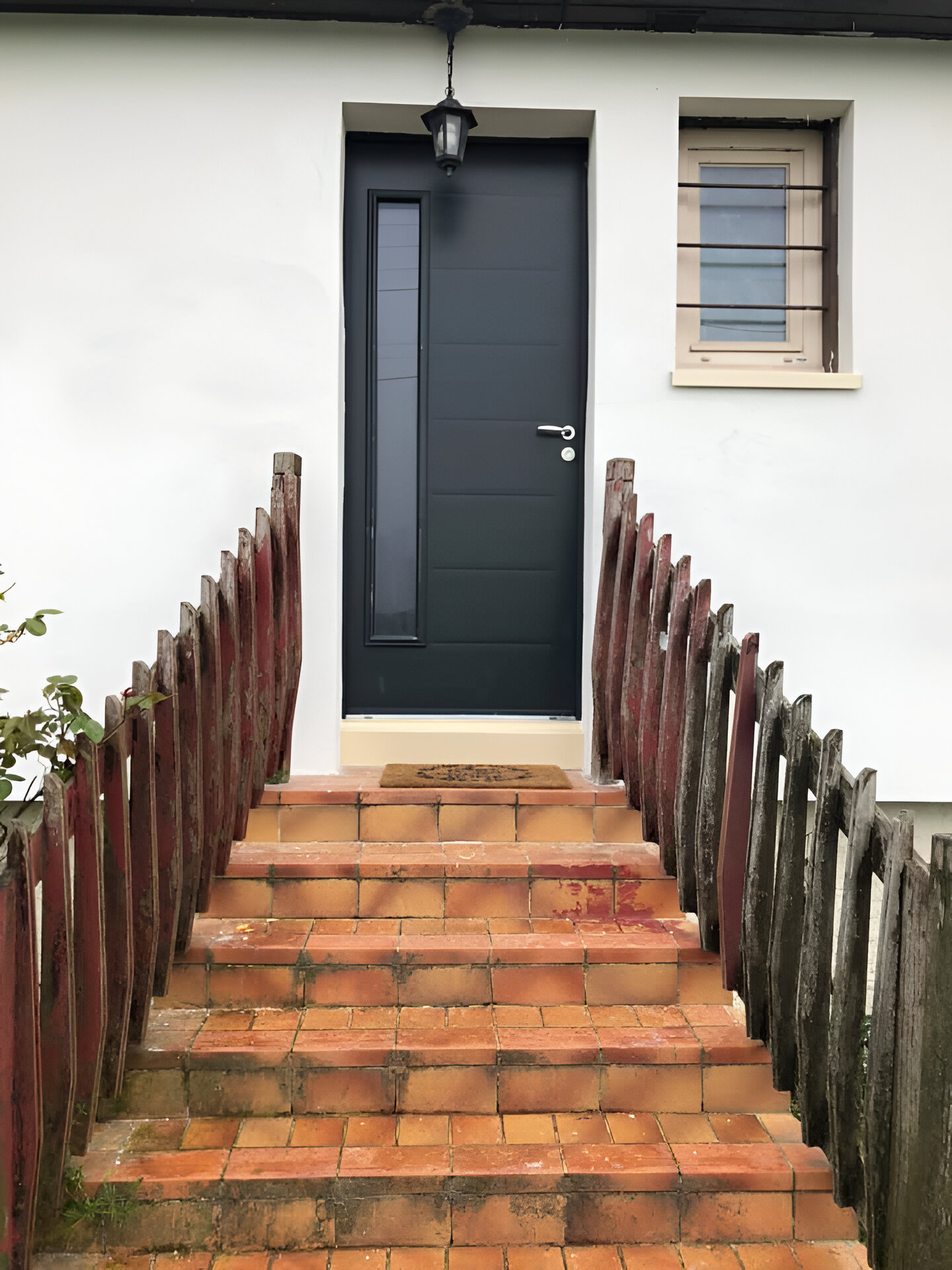 Des marches en briques mènent à une porte grise avec fenêtre latérale.