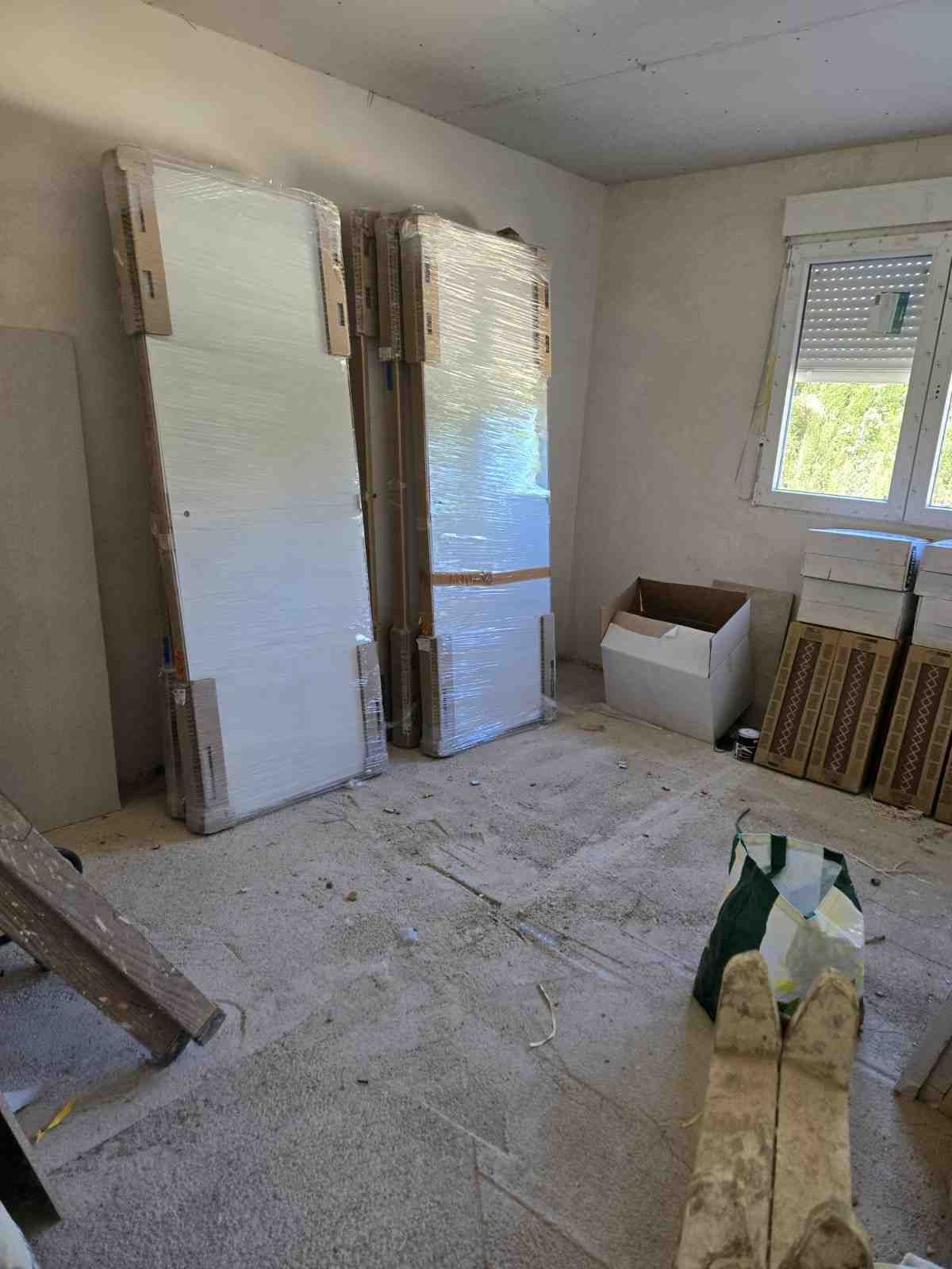 Habitación en obras con paredes sin terminar, tablas apiladas y materiales de construcción junto a dos puertas abiertas.
