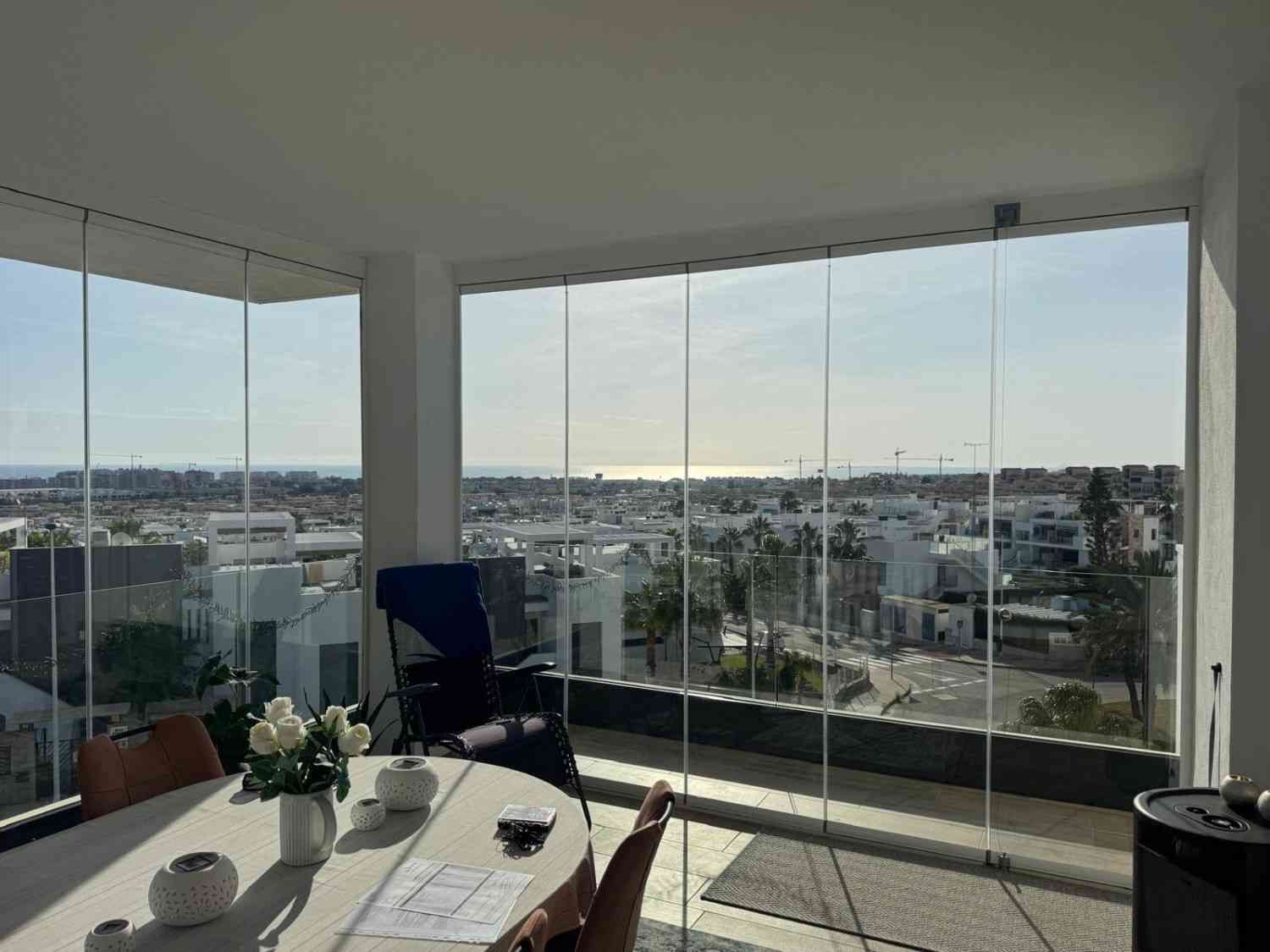 Balcón en esquina con barandilla de cristal, mesa y sillas de exterior, con vistas a la ciudad y al mar al atardecer.