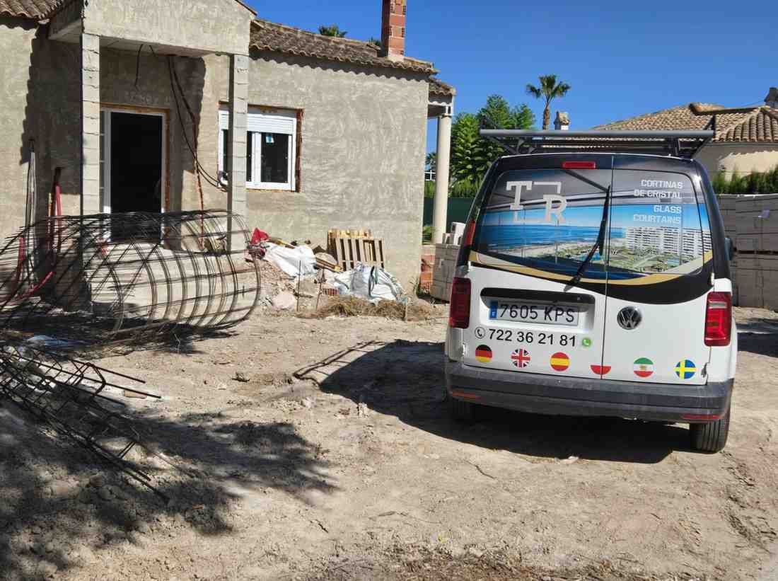 Furgoneta blanca aparcada junto a una pequeña casa de piedra en un patio polvoriento bajo un cielo azul despejado.