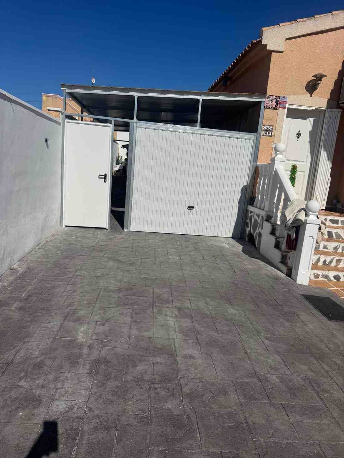 Entrada para vehículos con portón blanco y puerta lateral, suelo pavimentado y una pared de la casa.