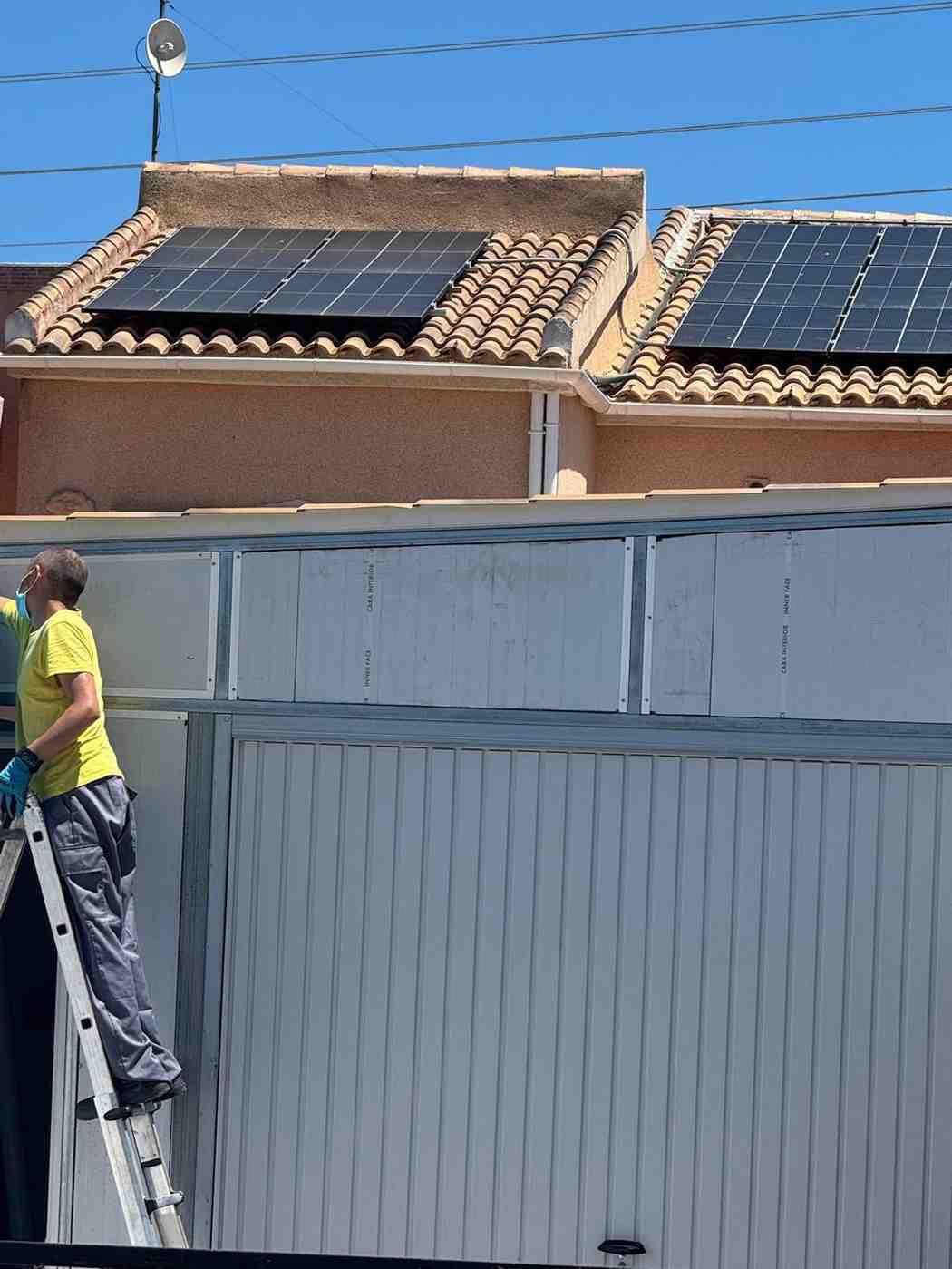 Un trabajador subido a una escalera junto a un tejado con paneles solares y una valla alta bajo un cielo despejado.