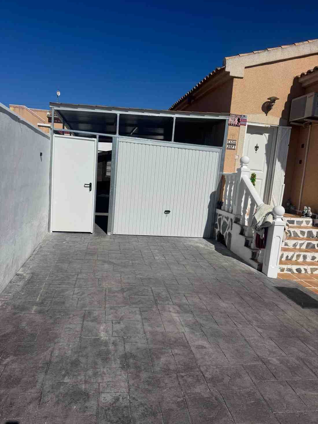Una puerta blanca abierta al final de un camino pavimentado junto a casas de color beige bajo un cielo azul claro.