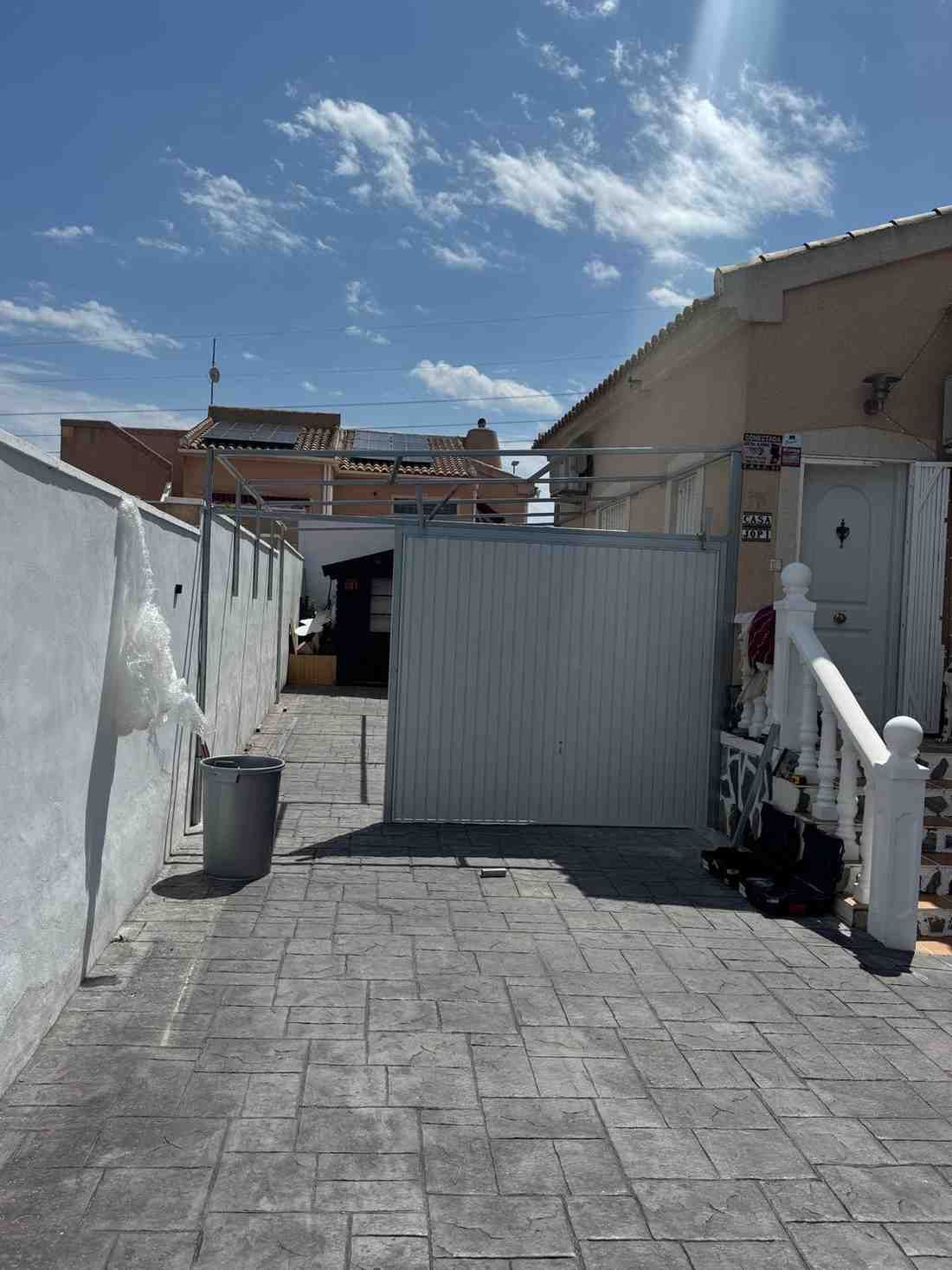 Un estrecho patio exterior con suelo de baldosas grises, paredes blancas y una puerta de metal corrugado bajo un cielo azul.