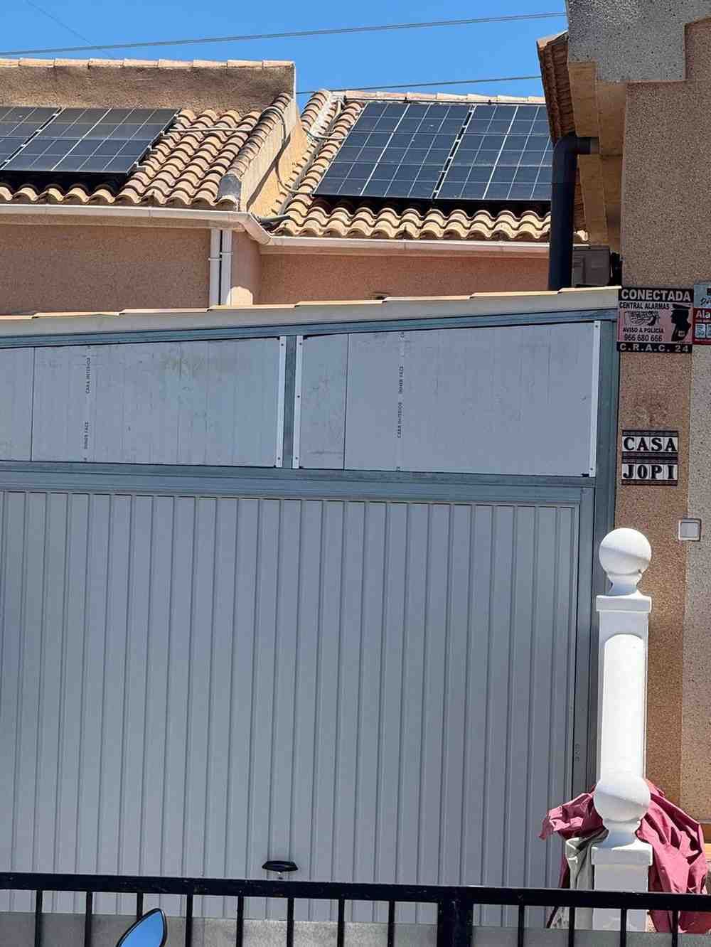 Puerta de garaje blanca y pared con tejado de tejas y paneles solares detrás, bajo un cielo azul despejado.
