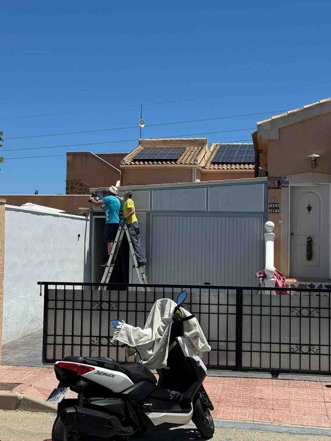 Un obrero, subido a una escalera, repara una puerta gris frente a una casa, con una moto aparcada en primer plano.