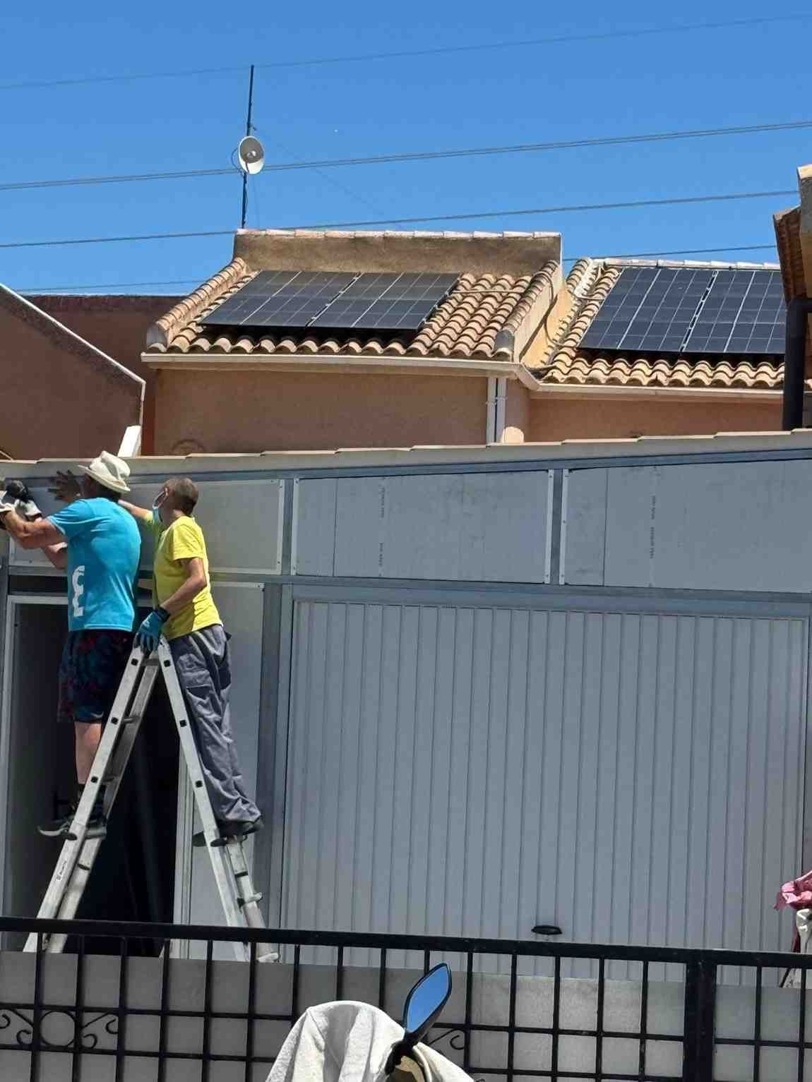 Dos trabajadores subidos a una escalera reparan un tejado sobre una valla blanca.