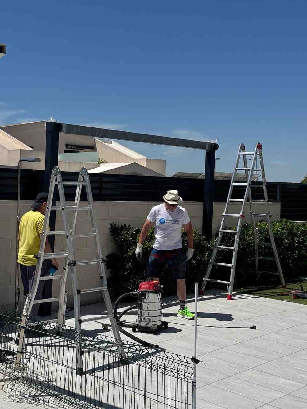 Dos personas pintando una pérgola en un patio trasero soleado, con escaleras y herramientas cerca.