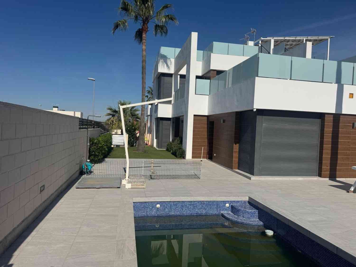Exterior de una casa moderna con piscina, patio y palmeras bajo un cielo azul despejado.