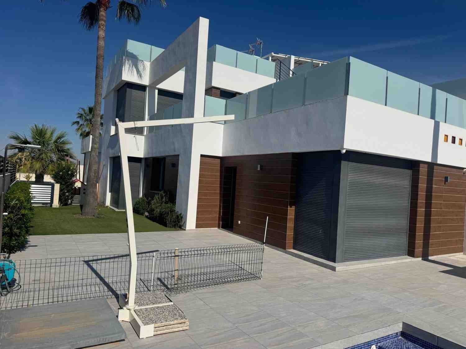 Casa moderna blanca y marrón con balcón, puerta de garaje y pequeño jardín delantero bajo un cielo azul despejado.