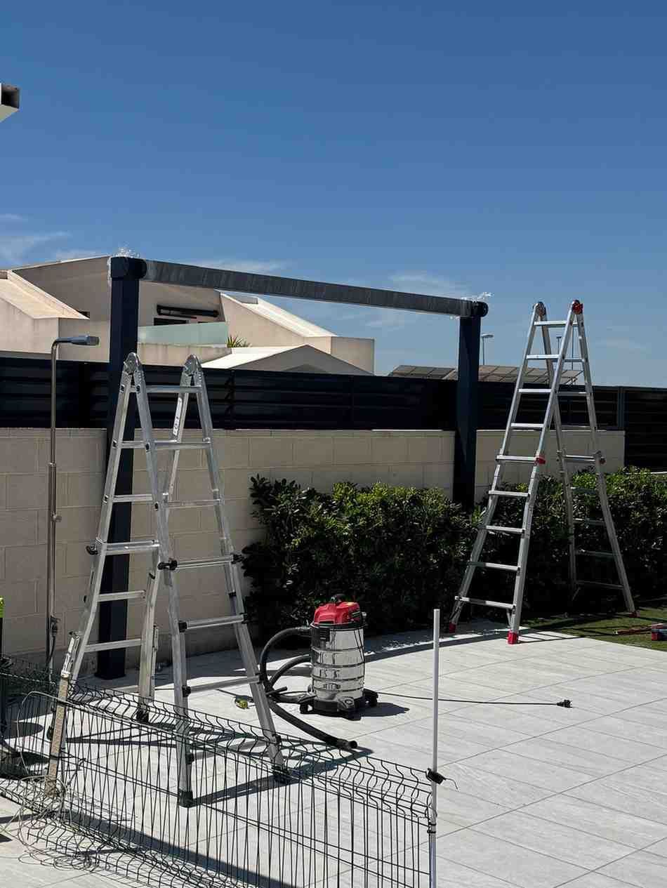Dos escaleras y un trabajador en una terraza en construcción, con una herramienta eléctrica en el suelo.
