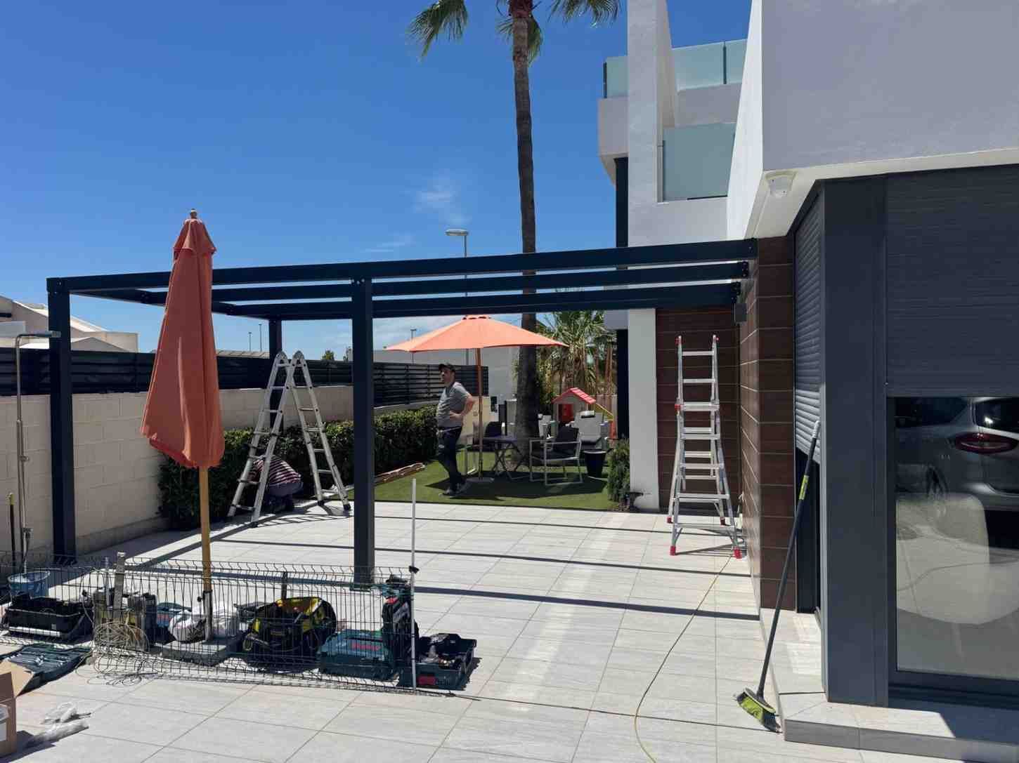 Patio trasero con pérgola, escaleras, sombrillas y un coche aparcado junto a una casa blanca.